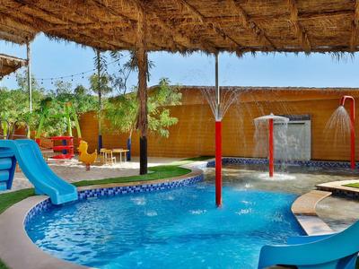 Área de juegos infantiles con tobogán y aspersores junto a una piscina bajo techo de paja.