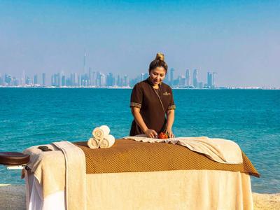 Mujer prepara mesa de masaje con mar y ciudad al fondo.