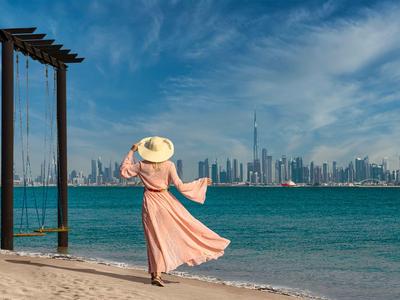 Woman in a pink dress and hat on the beach looking at a modern city skyline.