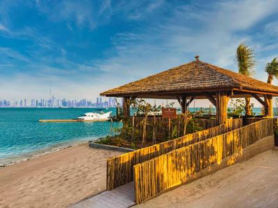 Beach with wooden pavilion and distant city view by the sea under a blue sky.