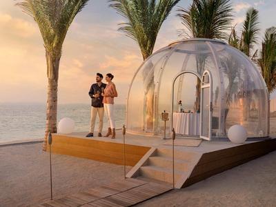 Two people stand beside a transparent dome on a platform at the beach during sunset.