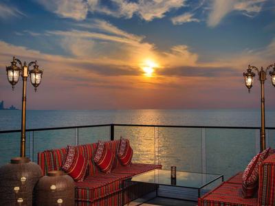 Cozy terrace with red-striped sofas and lanterns, sunset over the sea in the background.