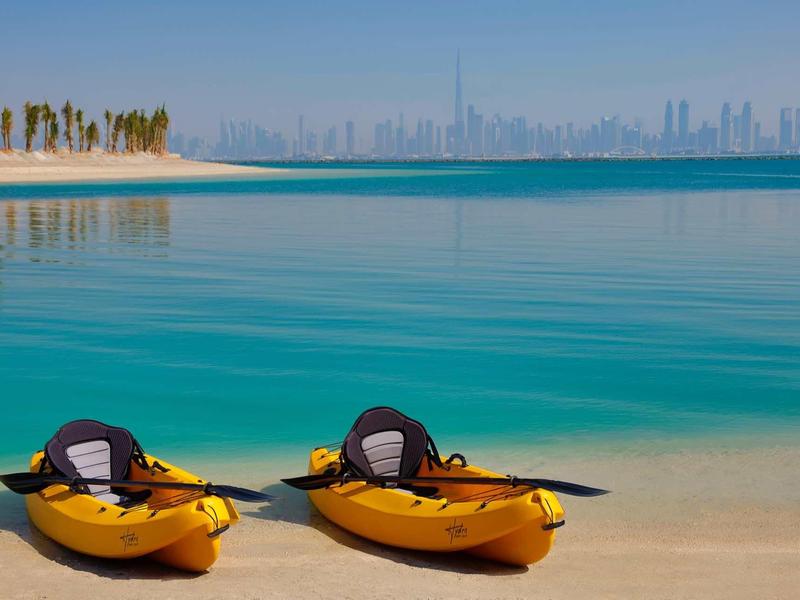 Dos kayaks amarillos en la orilla con agua turquesa y la ciudad de fondo bajo un cielo despejado.