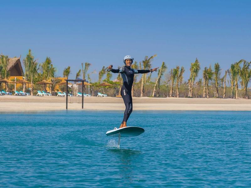 Persona sobre tabla de surf eléctrica en agua tranquila frente a playa con palmeras y cielo azul.