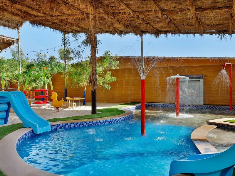 Área de juegos infantiles con tobogán y aspersores junto a una piscina bajo techo de paja.