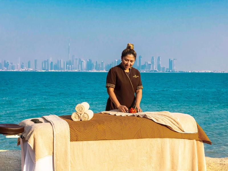 Mujer prepara mesa de masaje con mar y ciudad al fondo.
