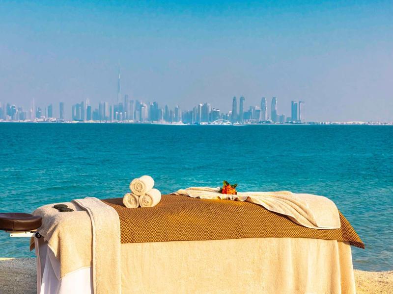 Camilla de masaje en la playa con vista al mar azul y el skyline a lo lejos.