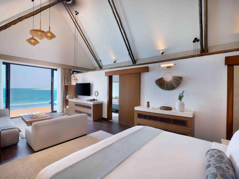 Habitación de hotel moderna con vista al mar, cama grande, área de sofá y TV bajo el techo inclinado.