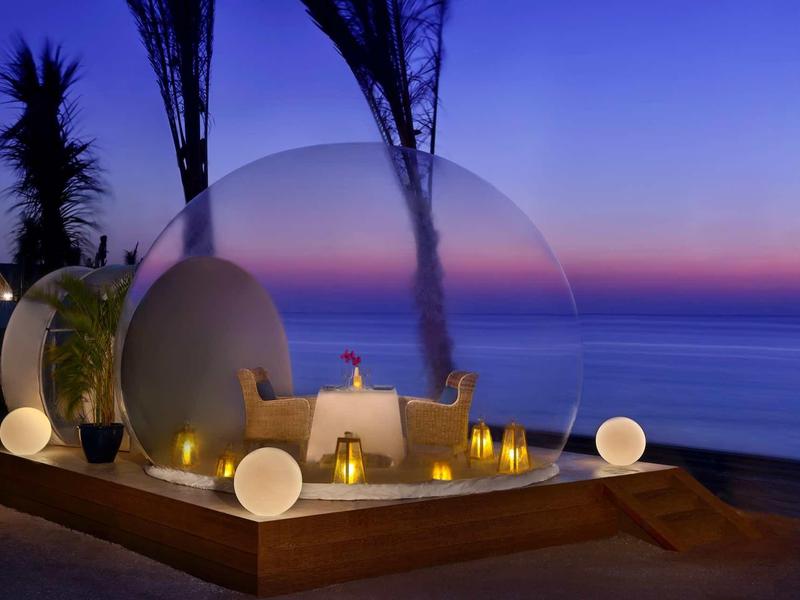 Cena romántica bajo una cúpula transparente con vista al mar al atardecer.