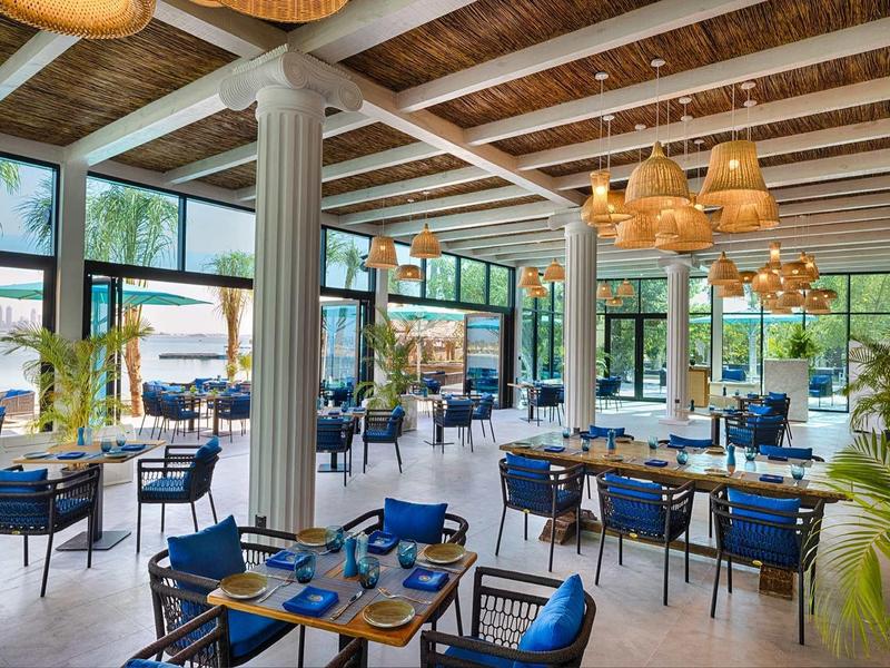 Restaurante luminoso con techo de vigas de madera, sillas azules y mesas preparadas en un hotel tropical.