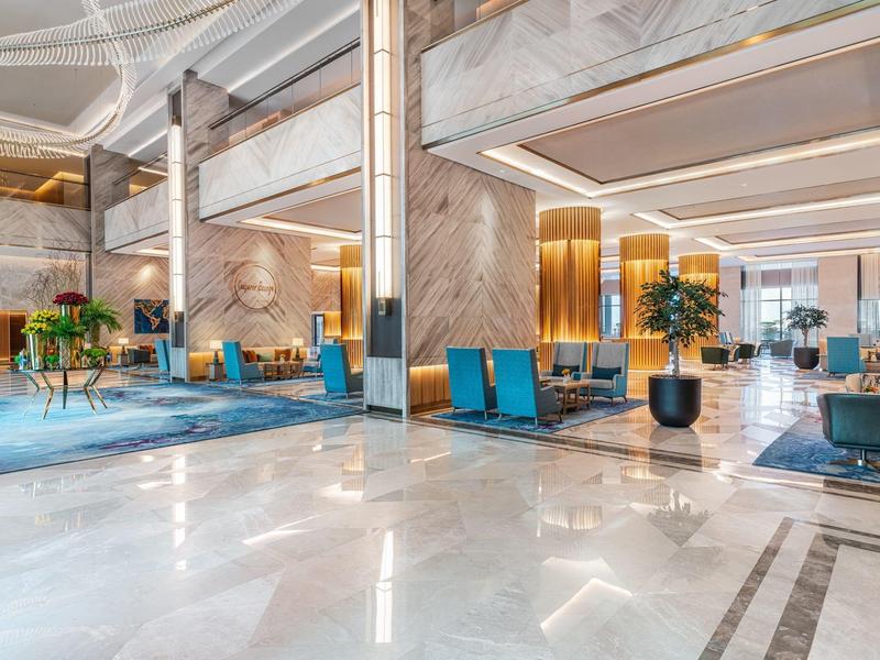 Große, helle Hotellobby mit Marmorfußboden, goldenen Akzenten, blauen Sesseln und grünen Pflanzen.
