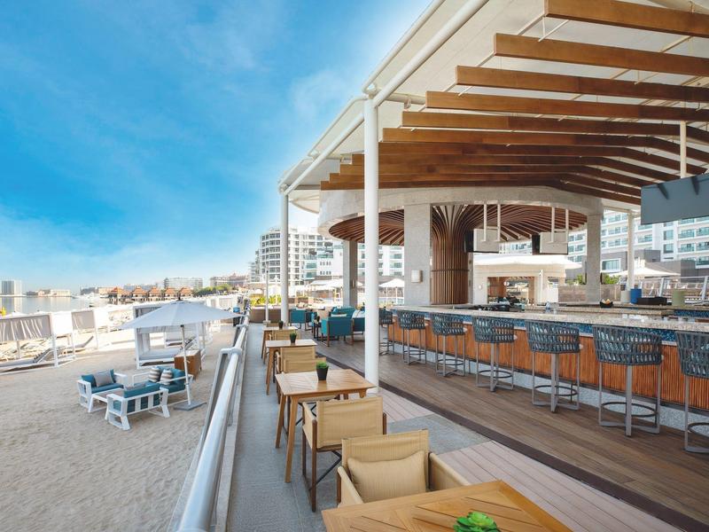 Offene Strandbar mit Holzmöbeln, Barhockern, blauen Sesseln und weitem Blick auf Sand und Meer.