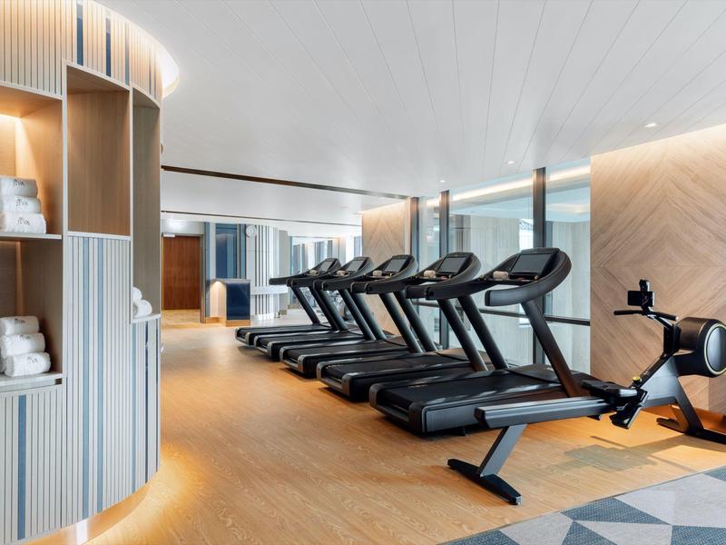 Moderner Fitnessraum mit Laufbändern, Rudergerät, Holzwänden und hellen LED-Leisten an der Decke.