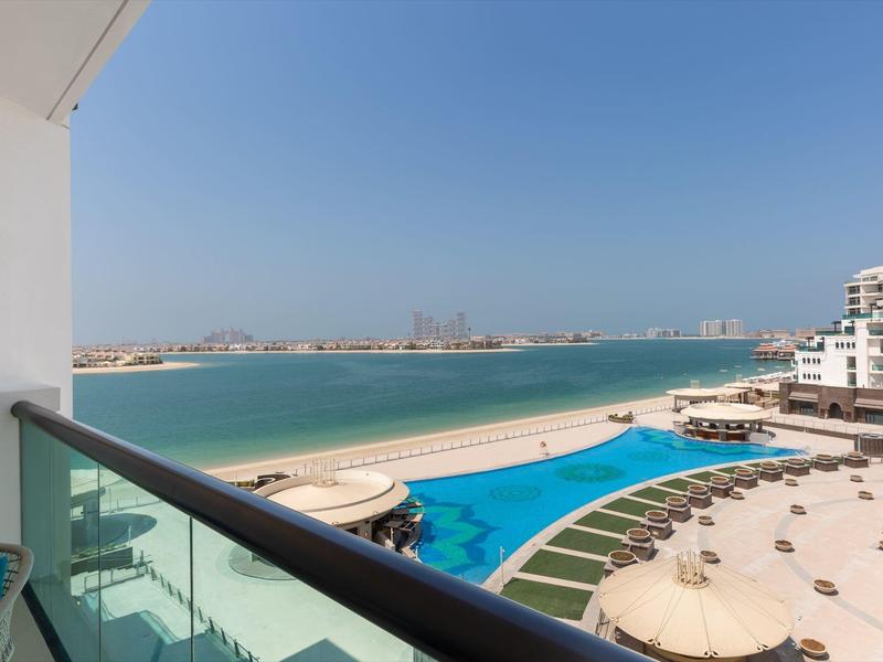 Meerblick von Balkon mit Glasgeländer auf Pool, Strand und moderne weiße Gebäude unter blauem Himmel.