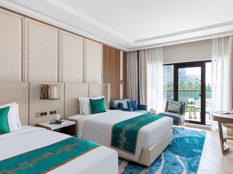 Hotelzimmer mit zwei Betten, blauen Teppich, Holzmöbeln und großem Fenster mit Balkon.