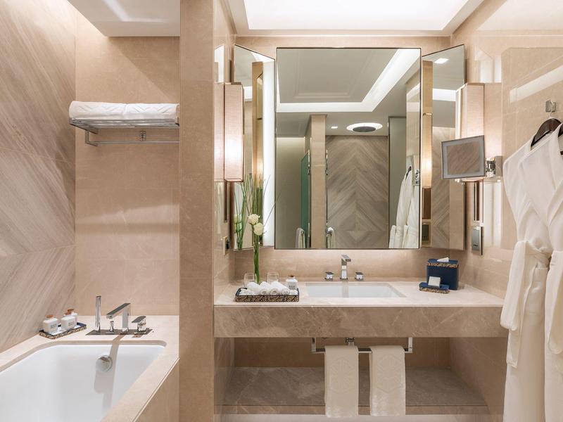 Modernes Badezimmer mit beiger Marmoroptik, Badewanne, großem Spiegel und weißen Bademänteln.