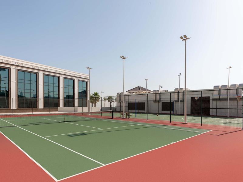 Ein leerer Tennisplatz mit grünem Spielfeld und rotem Rand, umgeben von Gebäuden und hellem Himmel.