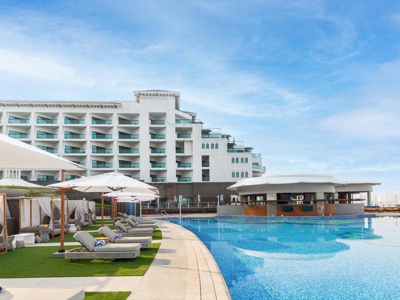 Großer Hotelpool mit Liegestühlen, Sonnenschirmen und einem mehrstöckigen Hotelgebäude im Hintergrund.