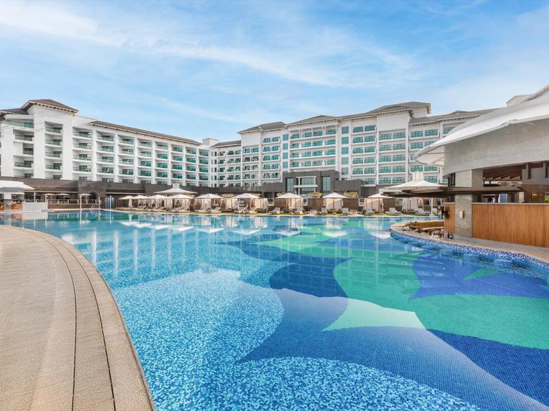 Ein großes Hotel mit hellen Zimmern um einen blauen Pool und Sonnenschirmen im Freien.