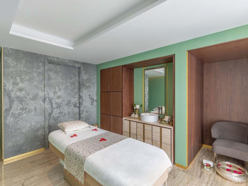 Modernes Spa-Zimmer mit Massageliege, grünen Wänden, grauem Sessel und Holz-Kommode.