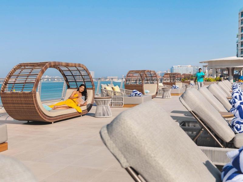 Strandliegen und runde Holzliegen am Pool mit Blick auf türkisfarbenes Meer unter blauem Himmel.