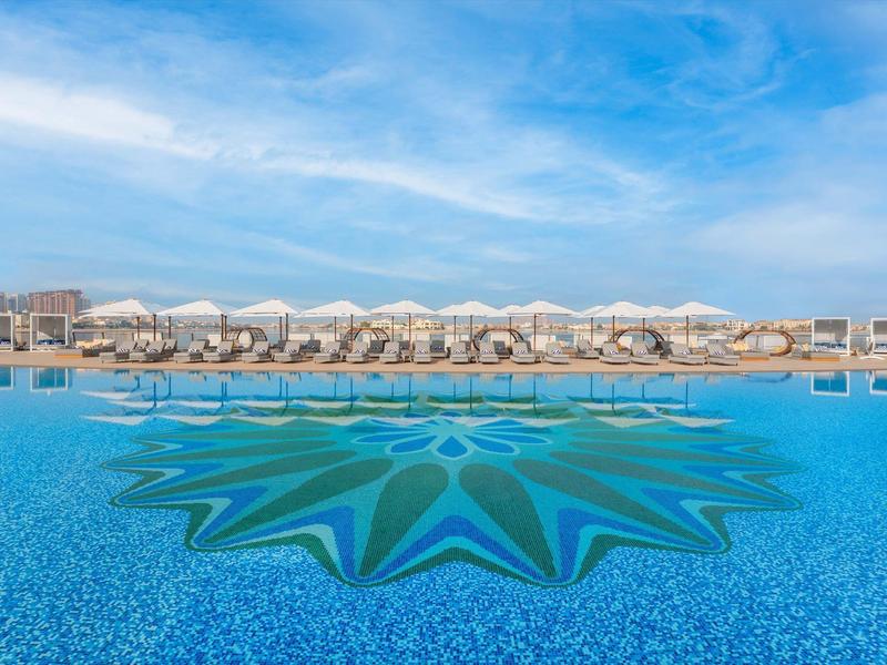 Großer Außenpool mit Sternmuster am Boden, Liegen und Sonnenschirmen, blauer Himmel mit Wolken.