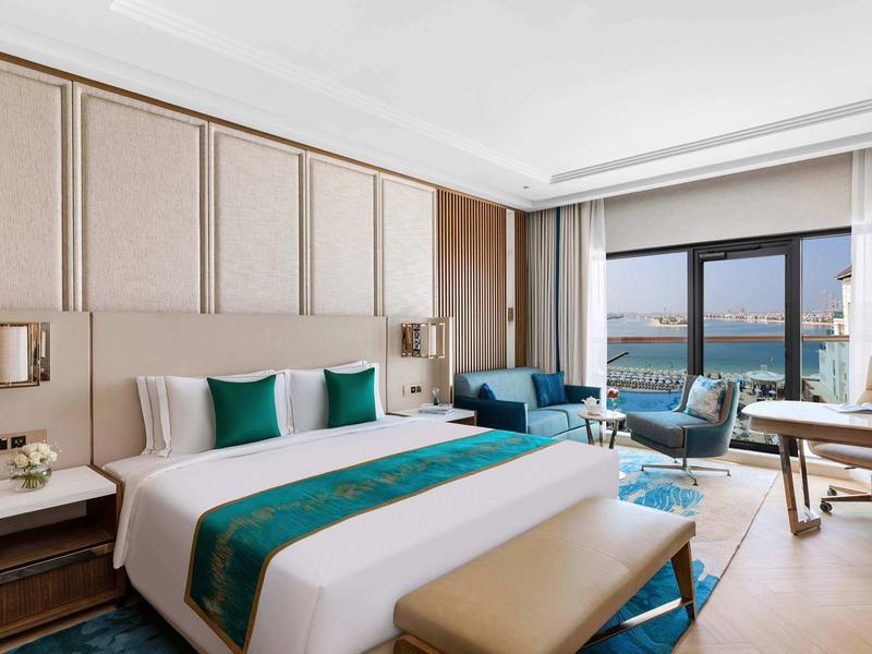 Geräumiges Hotelzimmer mit großem Bett, blau-grünen Akzenten, Sitzbereich und Blick auf das Meer.