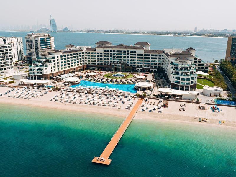Luxuriöses Strandresort mit langem Steg, Pool, Sandstrand und Meer in hellem Sonnenschein.