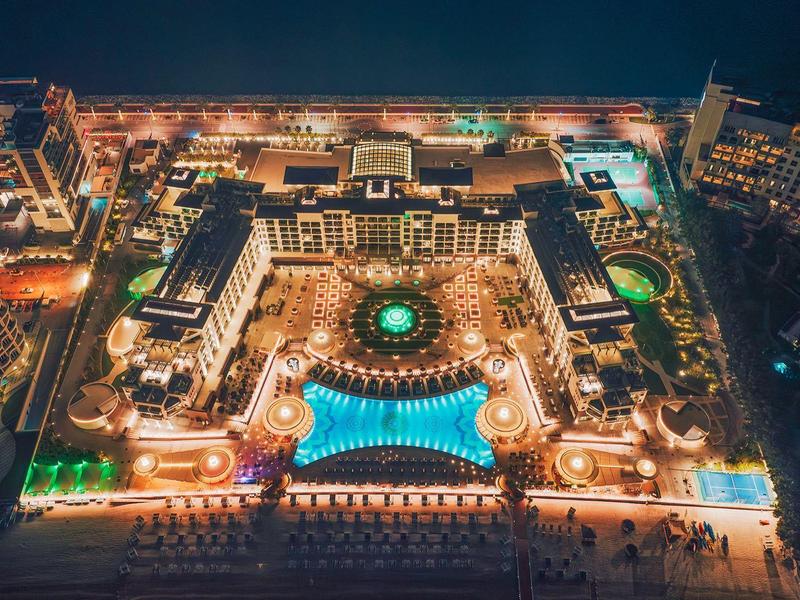 Nächtliche Luftaufnahme eines beleuchteten Hotels mit Pool, umgeben von Straßen und anderen Gebäuden.