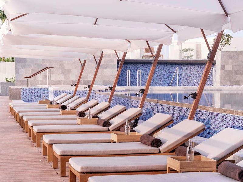 Reihe von weißen Liegestühlen mit Kissen unter Sonnenschirmen neben einem blauen Wasserbecken.