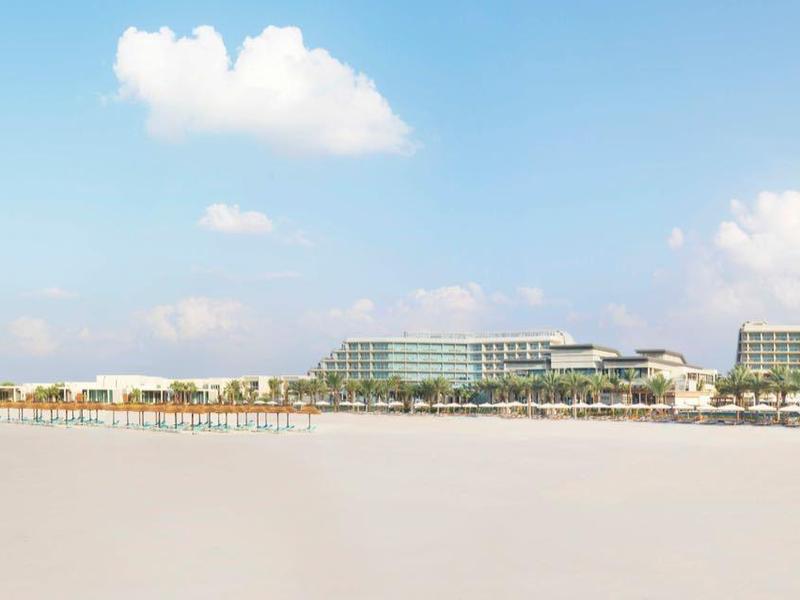Weitläufiger, heller Sandstrand mit blauem Meer, klarem Himmel und Gebäuden am Horizont.