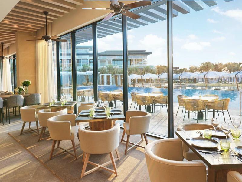 Modernes Restaurant mit Holzdecke, beigen Stühlen, Holzböden und großem Glasfenster zum Poolbereich.