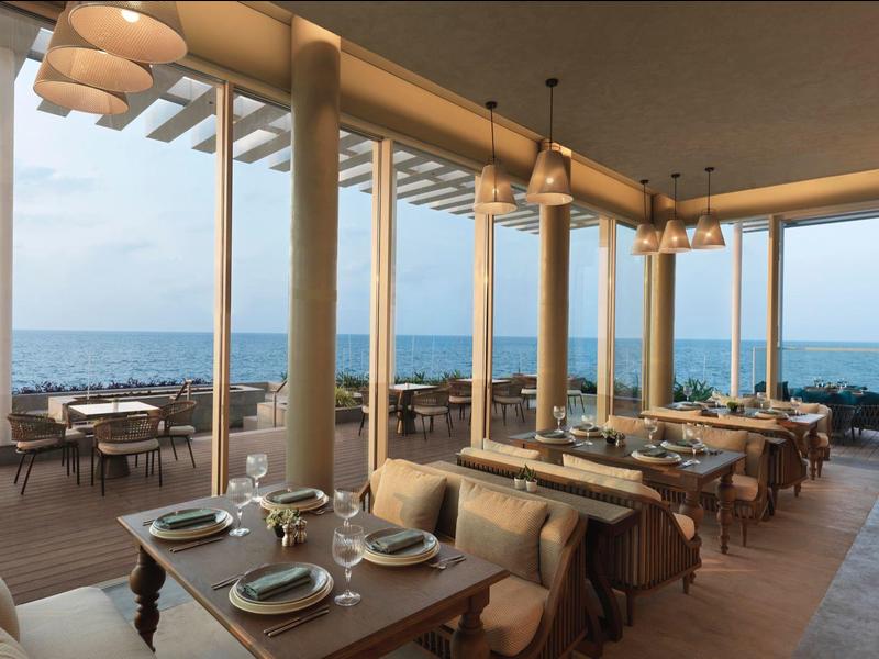 Restaurant mit großen Fenstern, Holztischen, Stühlen, Blick auf das offene Meer bei Tageslicht.