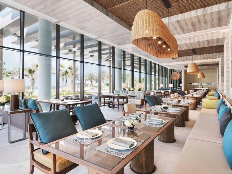 Modernes Restaurant mit großen Glasfenstern, Holzmöbeln und blauen Sitzkissen, helle und offene Atmosphäre.