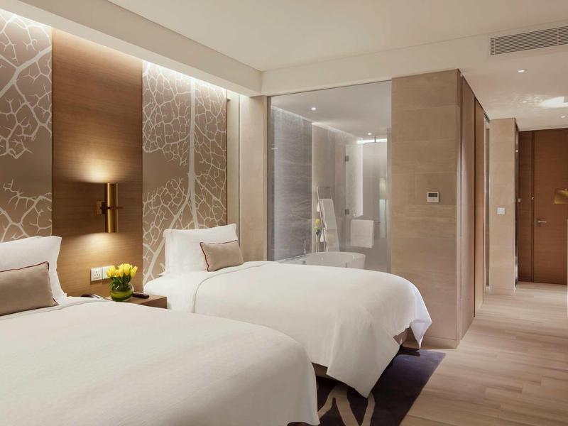Modern hotel room with two beds, indirect lighting, and an open bathroom area.