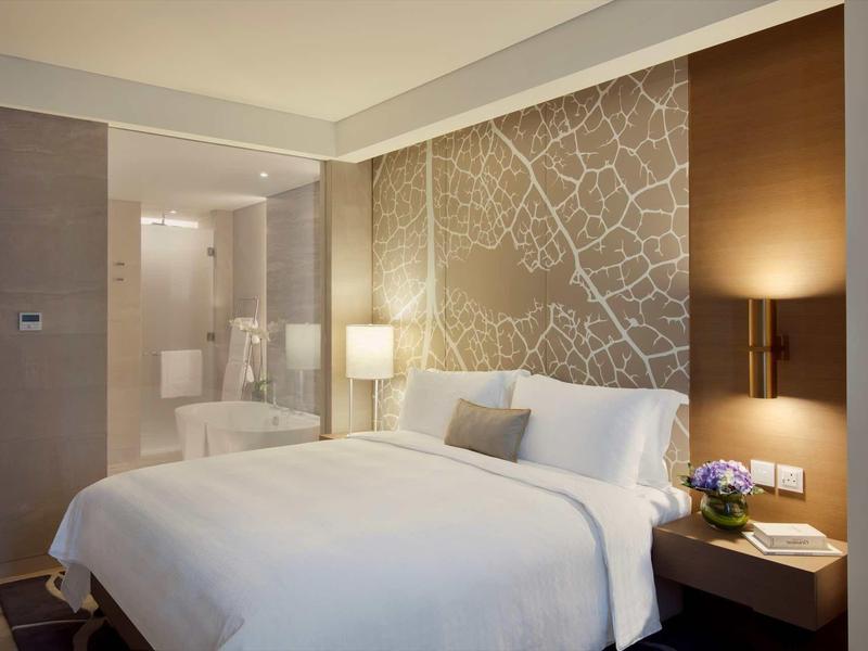 Modern hotel room with large bed, illuminated nightstand, and open bathroom area.
