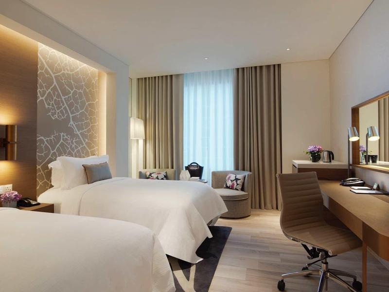 Modern hotel room with two single beds, desk, and window with light curtains.