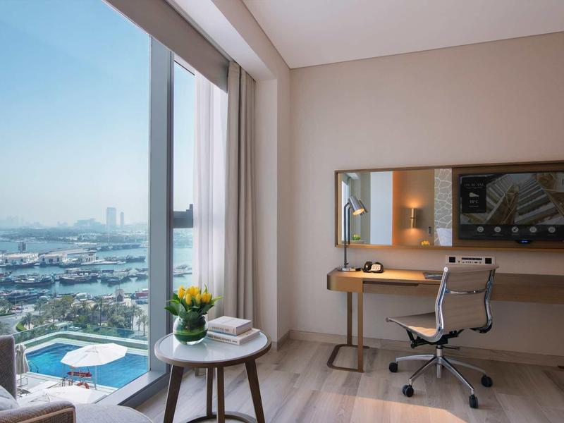 Bright hotel room corner with desk, chair, plants, and view of city and harbor.