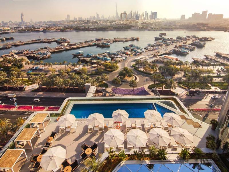 View of a hotel terrace with umbrellas and pool, with a harbor and city skyline in the background.