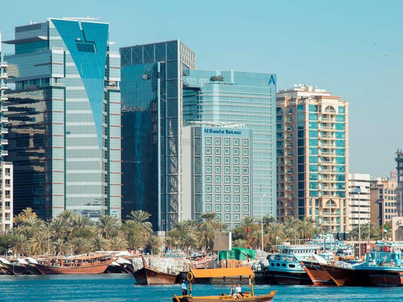 Modern high-rise buildings by the water with boats and palm trees under clear sky.