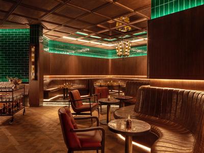 Gemütliche Bar mit braunen Ledersesseln, runden Tischen und grün-beleuchteten Akzenten.