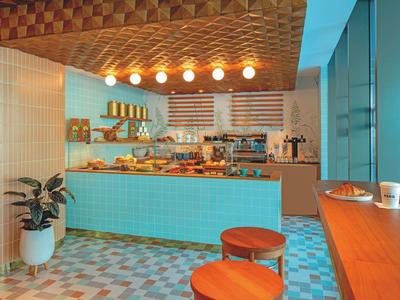 Moderne Café-Theke mit blauen Fliesen, Holzdecke, runden Hockern und großer Pflanze am Rand.