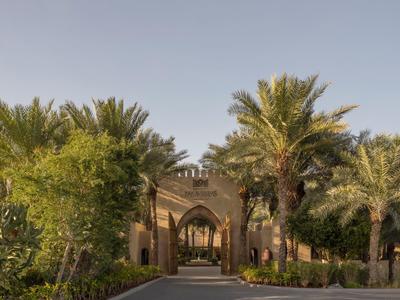 Hotel entrance with palm trees and greenery under clear blue sky.