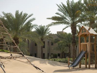Children's playground with slide and climbing net surrounded by palm trees in a hotel area.