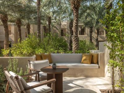 Cozy outdoor lounge with bench, chairs, and table surrounded by palm trees and green plants.