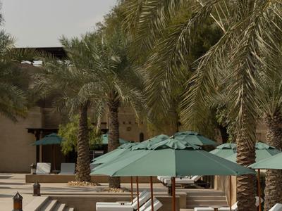 White lounge chairs under green umbrellas next to palm trees on a hotel terrace.
