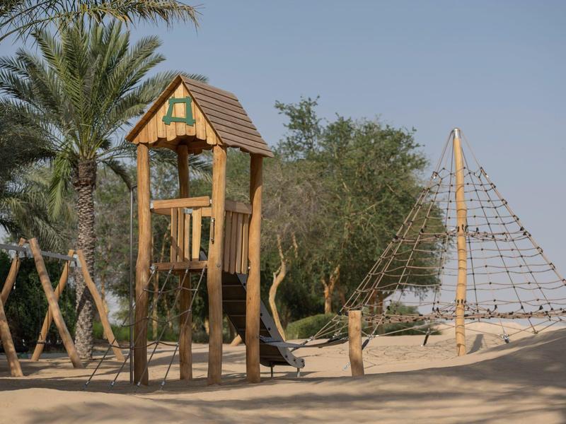 Sandy playground with wooden climbing structure and palm trees in the background.