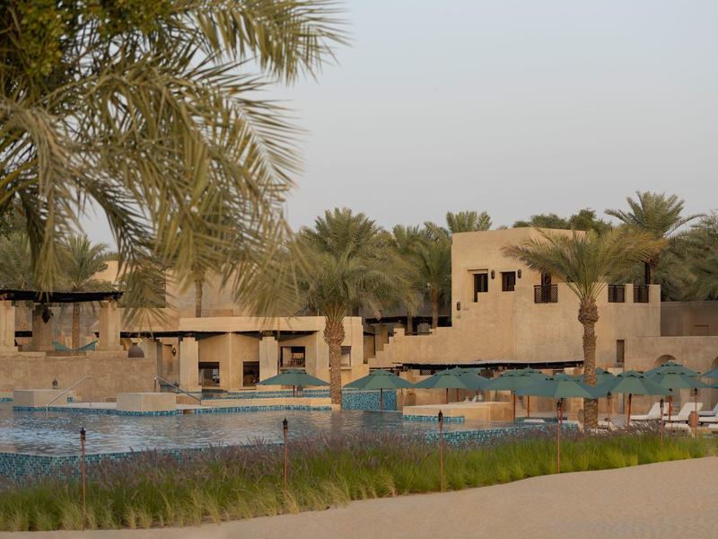 Hotel pool with lounge chairs and buildings in a sunny desert setting with palm trees.