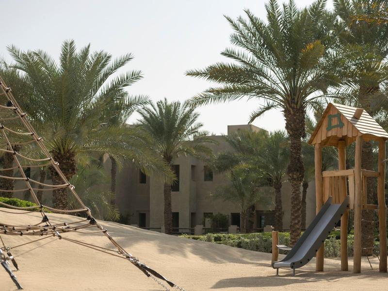 Children's playground with slide and climbing net surrounded by palm trees in a hotel area.