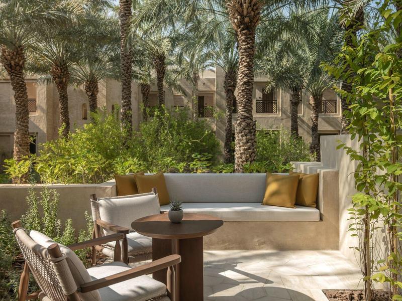 Cozy outdoor lounge with bench, chairs, and table surrounded by palm trees and green plants.